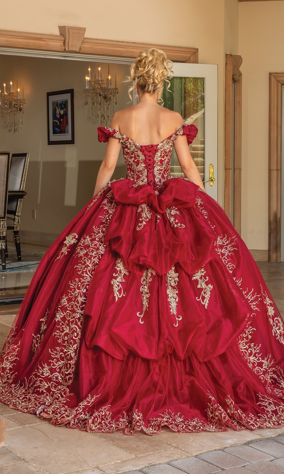 A person stands indoors wearing the Quinceanera Dress 1697 with Embroidery, a red off-the-shoulder gown featuring gold embroidery, layered ruffles, and a stunning sweetheart neckline, viewed from the back.